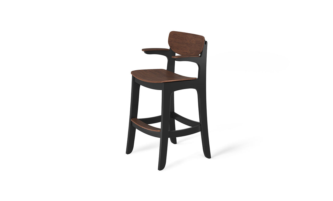 The Haven Hightop Chair by Ledge Lounger features a dark wooden seat and backrest on a sleek black frame. It offers ergonomic comfort with a footrest and one-sided armrest, showcasing a minimalist, contemporary design perfect for any setting.