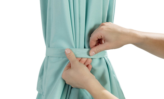 Close-up of hands tying a knot with a fabric belt around a light teal curtain, resembling fade-resistant canvas by Ledge Lounger. The plain white background highlights the texture and craftsmanship reminiscent of their 6.5 Square Signature 360° Tilt Umbrella.