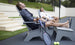 Two people relax on the Mainstay Ultimate Adirondack lounge chairs by Ledge Lounger under a clear sky. One holds a bottled drink, while a tennis racket and balls lie nearby, enjoying the serene surroundings and stylish poolside furniture.