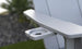 Close-up of the Mainstay Ultimate Adirondack by Ledge Lounger, a light gray outdoor lounge chair with a built-in cup holder. The blurred background of greenery and stones creates the perfect poolside relaxation setting.