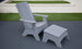 A Ledge Lounger Mainstay Adirondack Ottoman and matching chair sit on the dark patio. In the background, lush green grass borders a short white wall, creating a serene setting with high-quality materials.