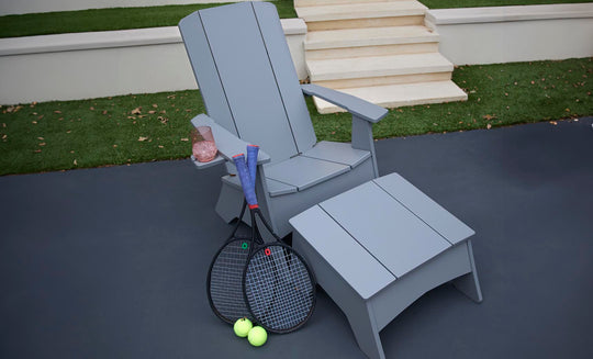 A gray Ledge Lounger chair features a matching Mainstay Adirondack Ottoman on the outdoor patio. Beside it lie two tennis rackets and balls, with a rolled-up blue towel over the arm next to a cup of pink drink, all set against steps and lush grass.