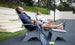 Two men relax on Ledge Lounger patio chairs with tennis rackets and balls nearby. One sips a drink as they both bask in the sun, eyes closed. Mainstay Adirondack Ottomans and steps leading to a grassy area add durability and style to the scene.