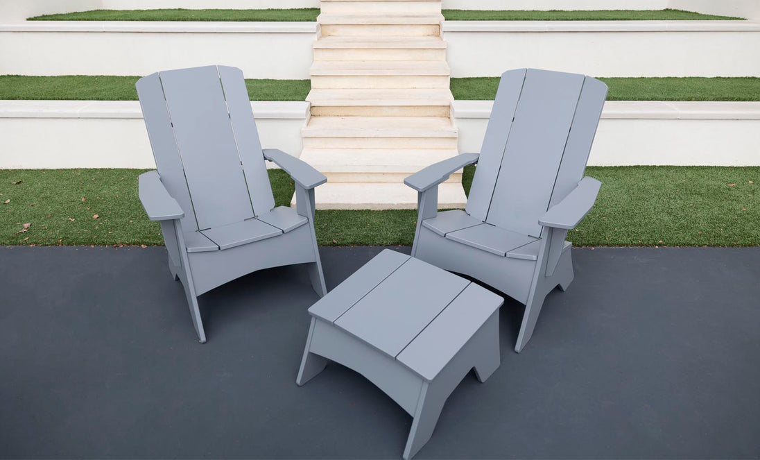 Two gray Ledge Lounger Adirondack chairs on a dark patio, showcasing durability and style, with a Mainstay Adirondack Ottoman in between. Background steps and grass patches complete the cozy outdoor setting.