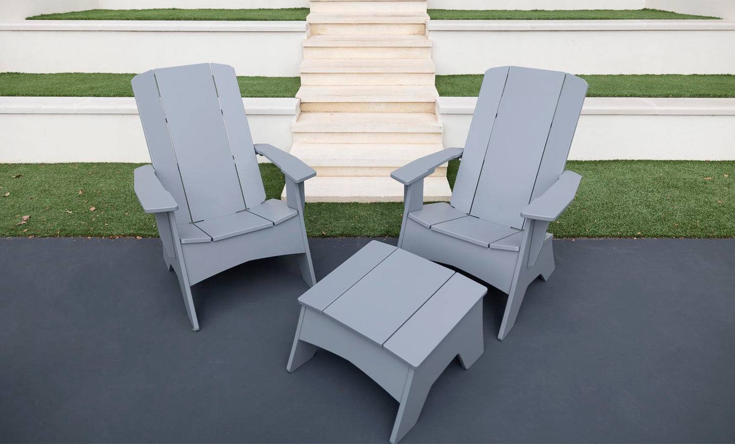 Two gray Ledge Lounger Adirondack chairs on a dark patio, showcasing durability and style, with a Mainstay Adirondack Ottoman in between. Background steps and grass patches complete the cozy outdoor setting.