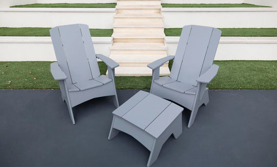 Two gray Ledge Lounger Adirondack chairs on a dark patio, showcasing durability and style, with a Mainstay Adirondack Ottoman in between. Background steps and grass patches complete the cozy outdoor setting.