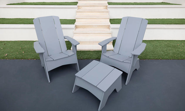Two gray Ledge Lounger Adirondack chairs on a dark patio, showcasing durability and style, with a Mainstay Adirondack Ottoman in between. Background steps and grass patches complete the cozy outdoor setting.