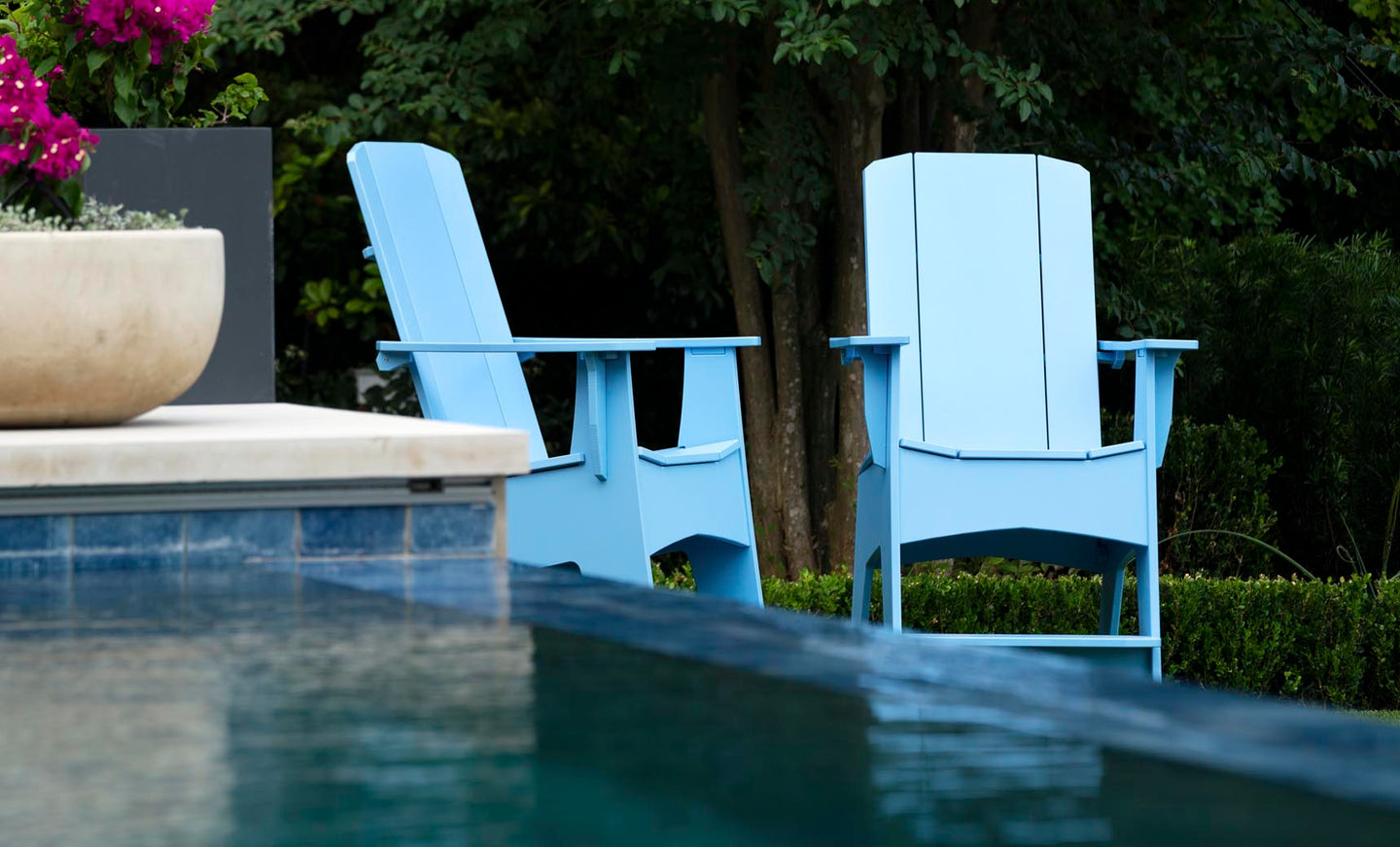Two Mainstay Adirondack Tall chairs from Ledge Lounger, in light blue and featuring built-in bottle openers, sit poolside amidst lush greenery. A planter of vibrant pink flowers on the left adds a pop of color to the tranquil outdoor setting.