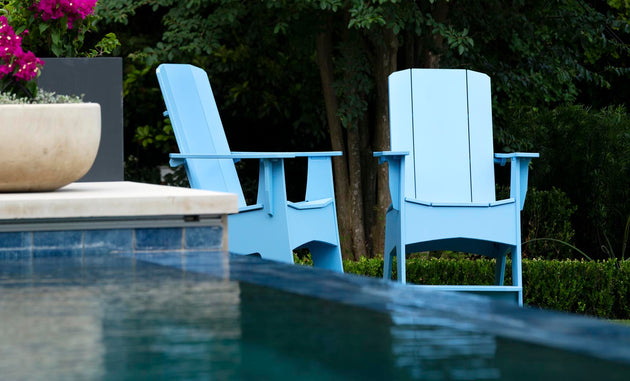 Two Mainstay Adirondack Tall chairs from Ledge Lounger, in light blue and featuring built-in bottle openers, sit poolside amidst lush greenery. A planter of vibrant pink flowers on the left adds a pop of color to the tranquil outdoor setting.
