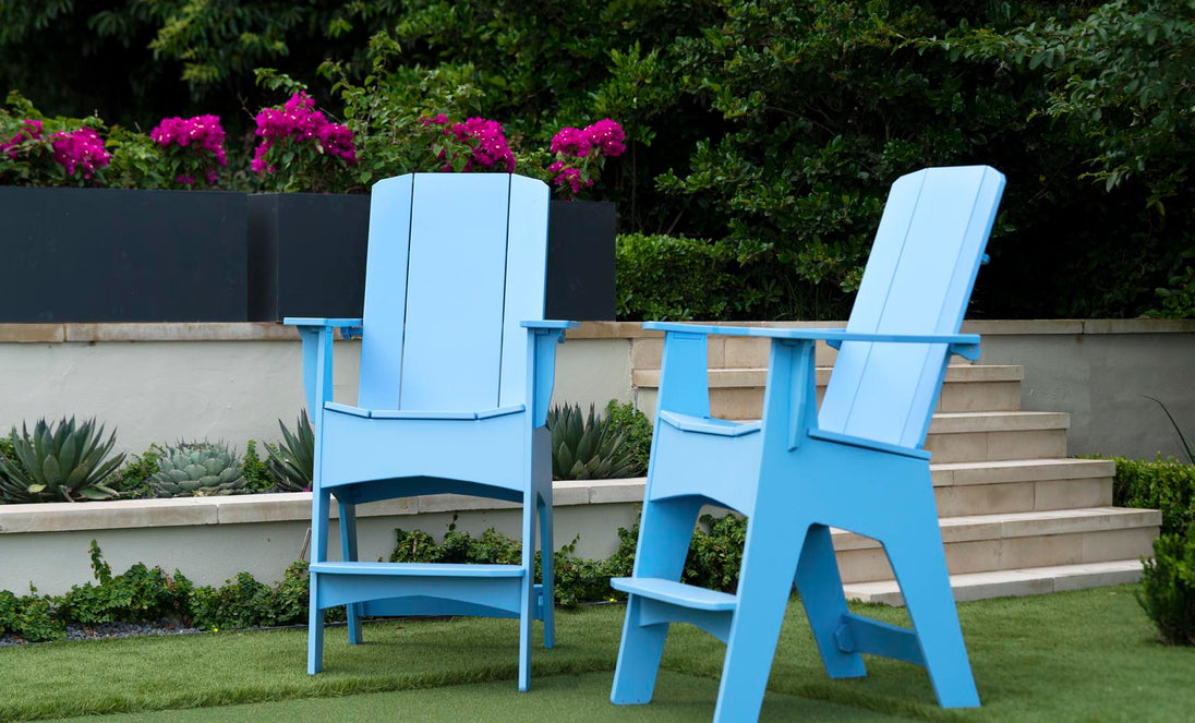 Two oversized Mainstay Adirondack Tall chairs by Ledge Lounger in light blue mark the ideal outdoor lounge setting. Set on the grass, concrete steps ascend behind with pink flowers flourishing. The vibrant garden scene is enhanced by built-in bottle openers for added convenience.