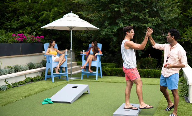 Two men exchange a high-five on the lawn near cornhole boards, while two women engage in conversation, relaxing in Ledge Loungers Mainstay Adirondack Tall chairs under a large umbrella. Theyre dressed in summer attire amid greenery, with a lounge seat featuring a built-in bottle opener nearby.