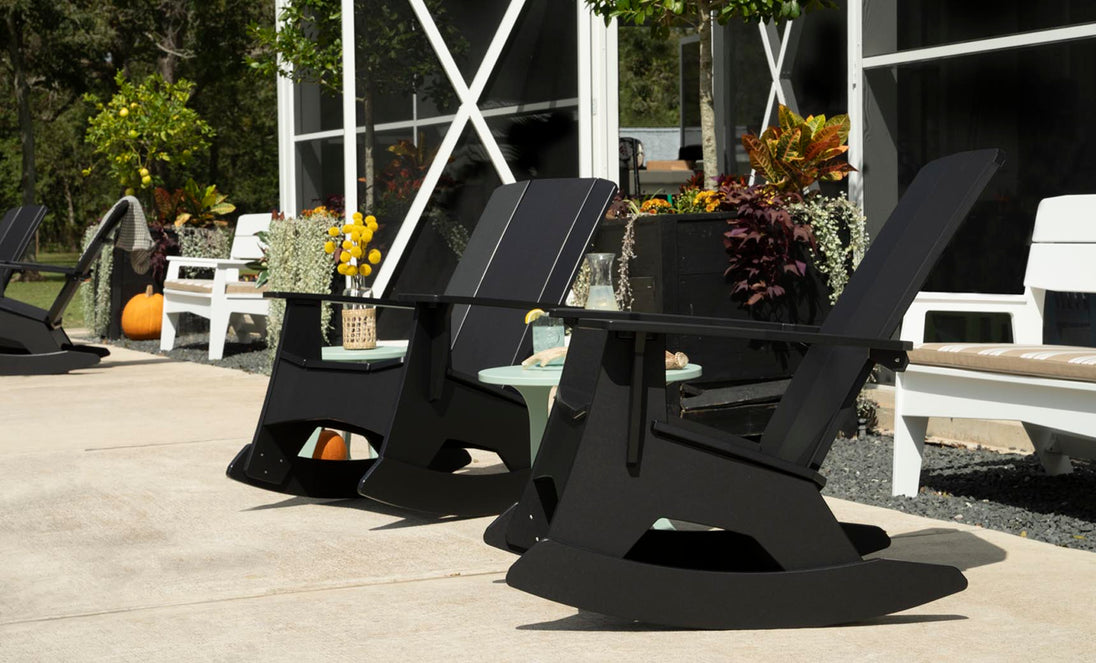 Mainstay Adirondack Rockers by Ledge Lounger enhance the sunny patio of this modern building. Enveloped by potted plants, pumpkins, and vivid foliage, its an ideal relaxation spot. A nearby table with yellow flowers in a decorative vase adds to the charming scene.