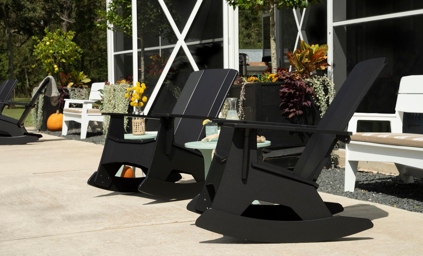 Mainstay Adirondack Rockers by Ledge Lounger enhance the sunny patio of this modern building. Enveloped by potted plants, pumpkins, and vivid foliage, its an ideal relaxation spot. A nearby table with yellow flowers in a decorative vase adds to the charming scene.