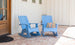 Two light blue Mainstay Adirondack Rockers by Ledge Lounger rest on a brick patio beside a wooden door, enhancing the serene space. One chair has a striped cushion. The white paneled wall with three small windows elegantly complements the setting.