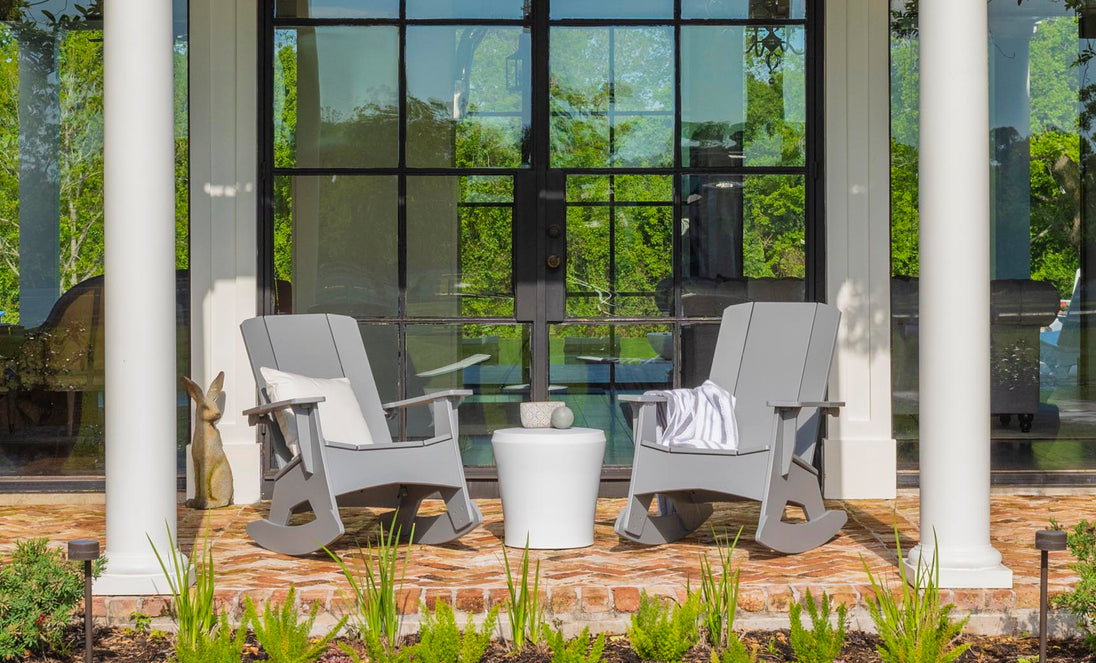 Two Mainstay Adirondack Rockers by Ledge Lounger, in gray, with a white side table between them sit on a brick patio. A striped blanket on one chair adds coziness. Large glass doors with lush greenery visible through the panes are in the background.