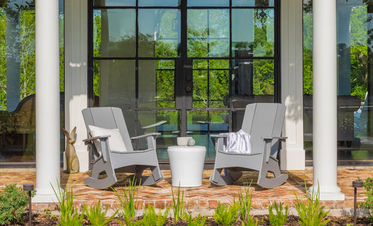 Two Mainstay Adirondack Rockers by Ledge Lounger, in gray, with a white side table between them sit on a brick patio. A striped blanket on one chair adds coziness. Large glass doors with lush greenery visible through the panes are in the background.