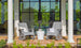Two Mainstay Adirondack Rockers by Ledge Lounger, in gray, with a white side table between them sit on a brick patio. A striped blanket on one chair adds coziness. Large glass doors with lush greenery visible through the panes are in the background.