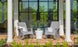 Two Mainstay Adirondack Rockers by Ledge Lounger, in gray, with a white side table between them sit on a brick patio. A striped blanket on one chair adds coziness. Large glass doors with lush greenery visible through the panes are in the background.