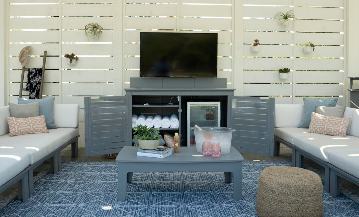 A cozy outdoor seating area features a large TV on the Mainstay Bar Credenza by Ledge Lounger. The credenza, with open doors revealing towels and a mini-fridge, complements two gray sofas with patterned cushions facing a table adorned with plants and a basket. Air plants enhance the wooden slat wall.