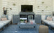 A cozy outdoor seating area features a large TV on the Mainstay Bar Credenza by Ledge Lounger. The credenza, with open doors revealing towels and a mini-fridge, complements two gray sofas with patterned cushions facing a table adorned with plants and a basket. Air plants enhance the wooden slat wall.