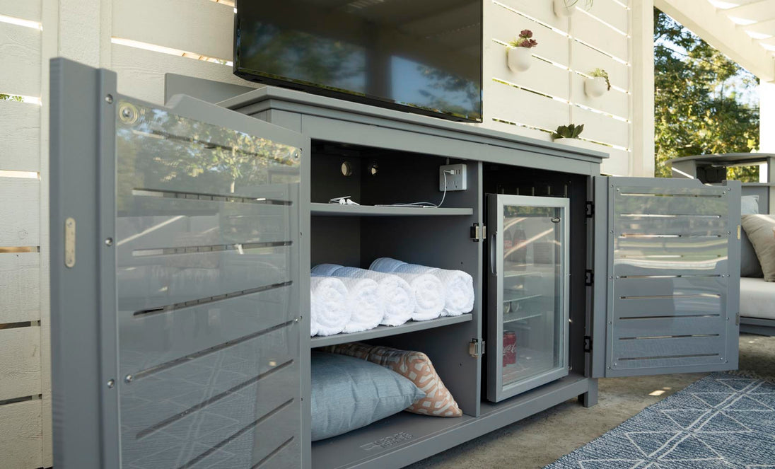 An outdoor entertainment haven boasts a TV above the Ledge Lounger Mainstay Bar Credenza, which offers ample storage. Open doors reveal stacked towels, a mini fridge, and a patterned cushion on the floor against a white slatted wall.