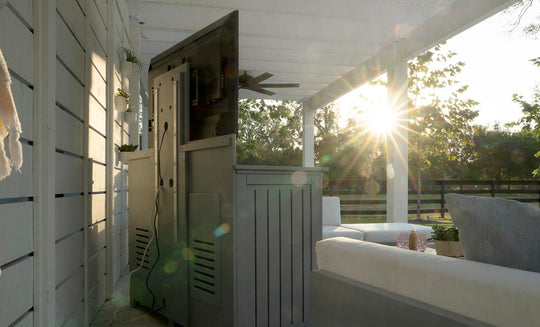 A sunlit outdoor patio features a large TV atop the Mainstay Bar Credenza - Final Sale by Ledge Lounger. White seating is partially sunlit, with trees in the background. Sun rays create lens flares, making the scene feel bright and inviting.