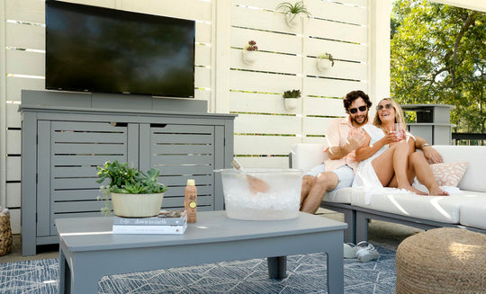 A couple relaxes on the patio, enjoying drinks on a Ledge Lounger gray outdoor sofa under a pergola with a TV. A Mainstay Bar Credenza holds their plant, magazine, and ice bucket as sunlight streams through the slatted walls.