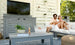 A couple relaxes on the patio, enjoying drinks on a Ledge Lounger gray outdoor sofa under a pergola with a TV. A Mainstay Bar Credenza holds their plant, magazine, and ice bucket as sunlight streams through the slatted walls.