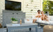 A couple relaxes on the patio, enjoying drinks on a Ledge Lounger gray outdoor sofa under a pergola with a TV. A Mainstay Bar Credenza holds their plant, magazine, and ice bucket as sunlight streams through the slatted walls.