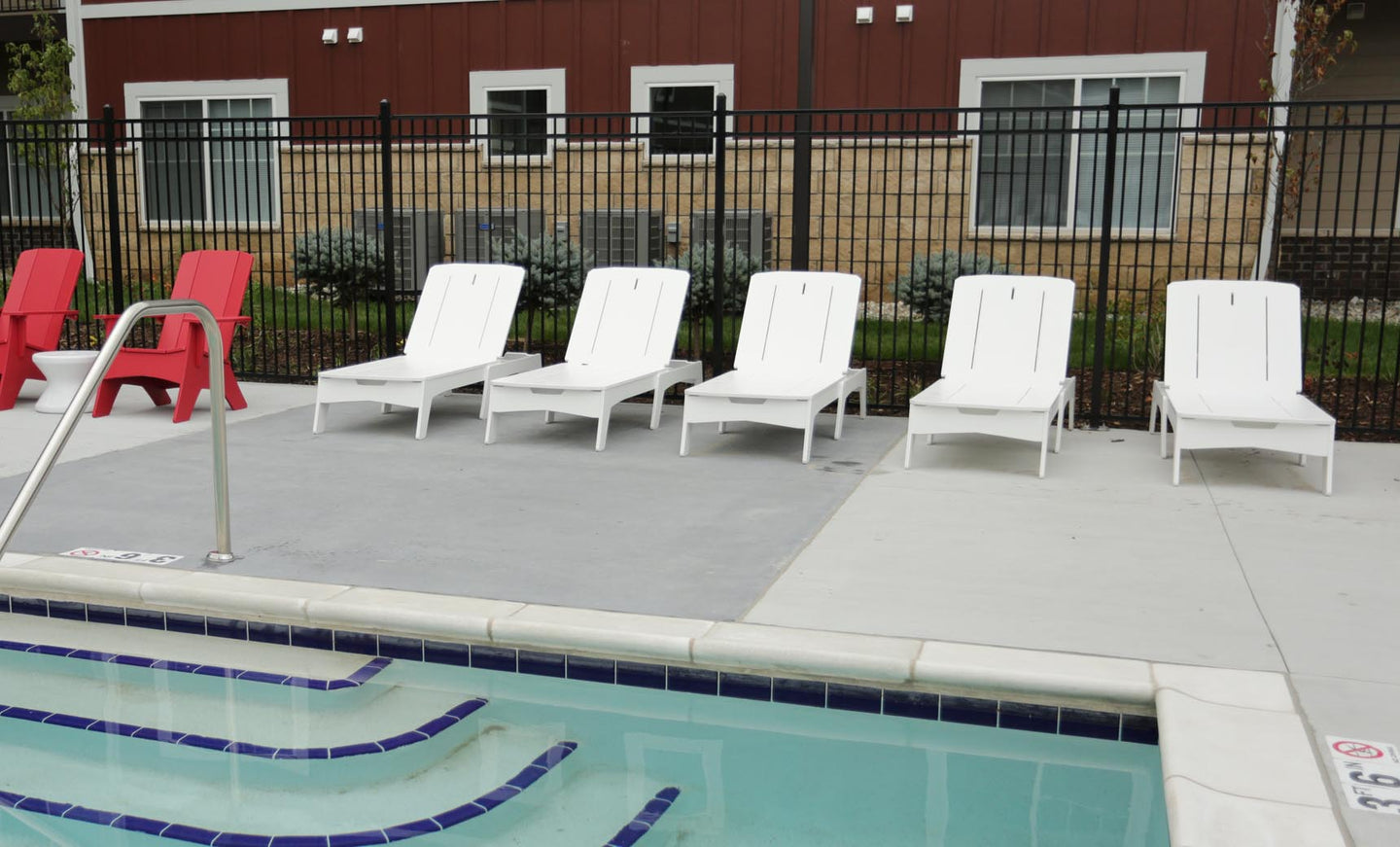 The outdoor pool area features five white Ledge Lounger Mainstay Chaises and two red chairs on a concrete deck. Stair railing leads into the pool with blue-bordered steps. A building with a two-tone exterior and windows is in the background, separated by a black fence.