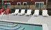 The outdoor pool area features five white Ledge Lounger Mainstay Chaises and two red chairs on a concrete deck. Stair railing leads into the pool with blue-bordered steps. A building with a two-tone exterior and windows is in the background, separated by a black fence.