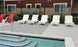 The outdoor pool area features five white Ledge Lounger Mainstay Chaises and two red chairs on a concrete deck. Stair railing leads into the pool with blue-bordered steps. A building with a two-tone exterior and windows is in the background, separated by a black fence.