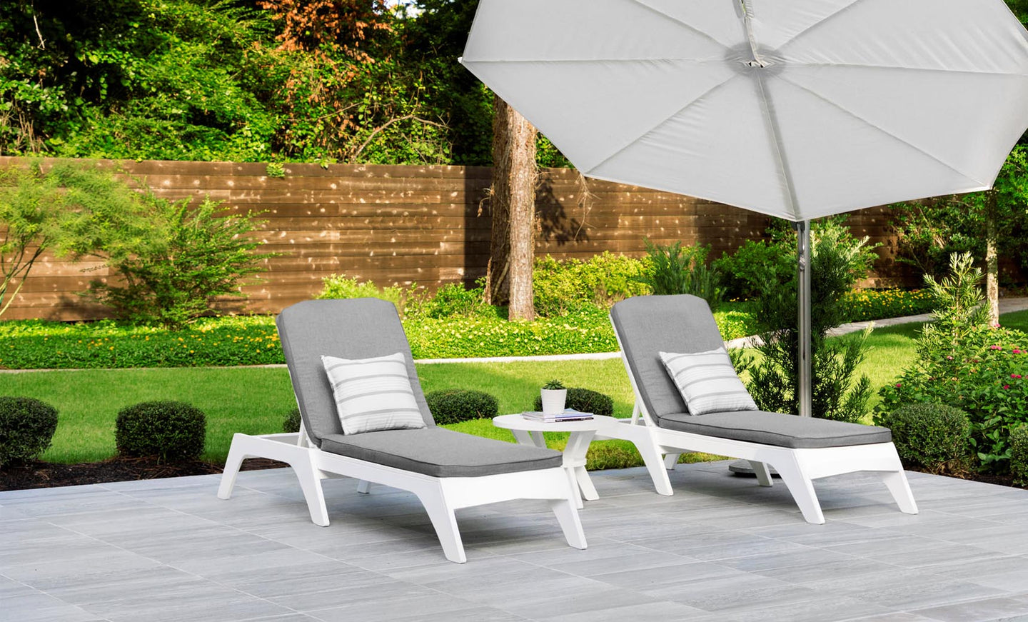 Two Ledge Lounger Mainstay Chaise lounge chairs, featuring gray cushions and striped pillows, are positioned on a tiled patio beneath a large white umbrella. A small round table with a cup enhances this versatile outdoor setup. A lush garden with a wooden fence serves as the background.
