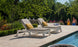 Two Ledge Lounger Mainstay Chaise lounge chairs with yellow cushions grace the stone deck. A couple in swimwear strolls by, while lush greenery enhances the serene ambiance. The pool edge glimmers invitingly in the foreground.