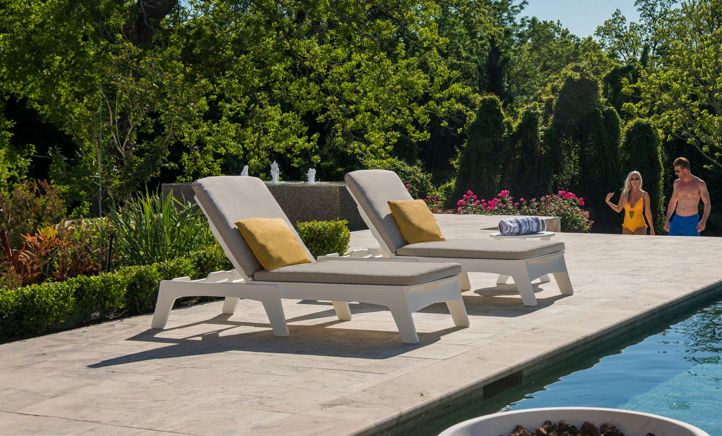 Two Ledge Lounger lounge chairs with Mainstay Chaise Cushions and yellow pillows sit on a stone poolside terrace, with a folded striped towel on one. In the background, greenery and blooming flowers frame a man and woman walking.