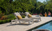 Two Ledge Lounger lounge chairs with Mainstay Chaise Cushions and yellow pillows sit on a stone poolside terrace, with a folded striped towel on one. In the background, greenery and blooming flowers frame a man and woman walking.