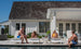 Four people enjoy a bright, sunny day by a modern white house with a wooden roof. Two chat and one reclines in full-body comfort on Mainstay Chaise Cushions from Ledge Lounger, while another sits at the pools edge in luxurious relaxation.