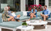 Four casually dressed people sit on an outdoor patio sofa surrounded by potted plants and red flowers, their lively conversation centered around a Mainstay Round Coffee Table by Ledge Lounger, enhancing the minimalistic and sunny setting.