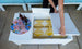 In an outdoor living space, a Mainstay Storage Coffee Table by Ledge Lounger with a white lid stores yellow-patterned blankets and supports a tray with three soda bottles and two glasses. Blue cushioned seating enhances the comfort of this inviting setting.