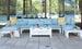 Outdoor living space with a light blue sectional sofa and yellow cushions. A Mainstay Storage Coffee Table by Ledge Lounger is centered, displaying bottles and a magazine. A straw hat, striped tote, and potted plants rest nearby. Large windows reflect the surrounding greenery.
