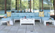 Outdoor living space with a light blue sectional sofa and yellow cushions. A Mainstay Storage Coffee Table by Ledge Lounger is centered, displaying bottles and a magazine. A straw hat, striped tote, and potted plants rest nearby. Large windows reflect the surrounding greenery.