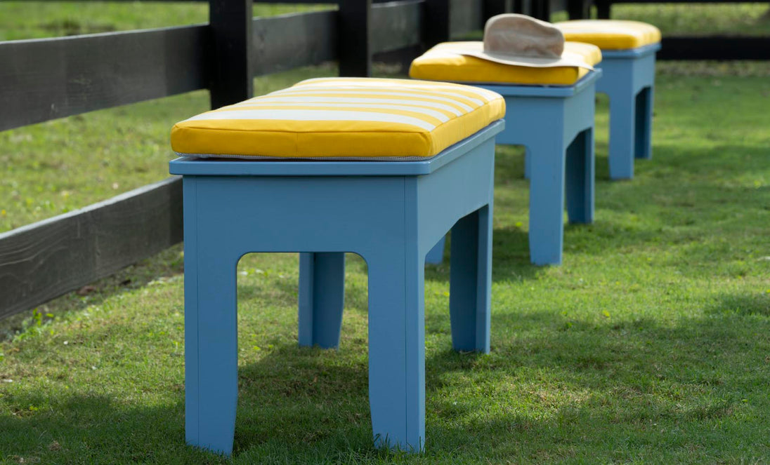 Three blue benches featuring Mainstay Bench Cushions by Ledge Lounger, with yellow stripes, line a grassy area next to a black fence. A beige hat sits on the back bench, enhancing the vibrant scene in this serene garden or park setting.