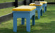 Three blue benches featuring Mainstay Bench Cushions by Ledge Lounger, with yellow stripes, line a grassy area next to a black fence. A beige hat sits on the back bench, enhancing the vibrant scene in this serene garden or park setting.