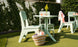Enjoy a blissful outdoor dining setup with Ledge Lounger’s Mainstay Dining Side Chairs and two light green tables on artificial grass. A woven basket with a magazine and pink water bottle sits below, while a pink cloth drapes over one chair. Yellow flowers add charm to this high-quality scene.