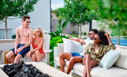 Four people enjoy a fire pit on Ledge Loungers Mainstay Sectional Relaxed Left Corner. Two chat on the bench, while the others relax in nearby chairs. Encircled by lush greenery, this serene spot offers endless configurations and comfort with a cushion set.