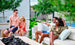 Four people enjoy a fire pit on Ledge Loungers Mainstay Sectional Relaxed Left Corner. Two chat on the bench, while the others relax in nearby chairs. Encircled by lush greenery, this serene spot offers endless configurations and comfort with a cushion set.