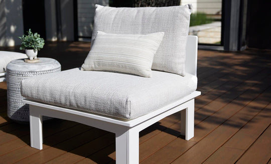 The Mainstay Sectional Relaxed Middle by Ledge Lounger is a white cushioned outdoor chair with a small striped pillow, part of a cozy set, on a wooden deck. A round side table beside it holds a green plant.