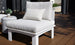 The Mainstay Sectional Relaxed Middle by Ledge Lounger is a white cushioned outdoor chair with a small striped pillow, part of a cozy set, on a wooden deck. A round side table beside it holds a green plant.