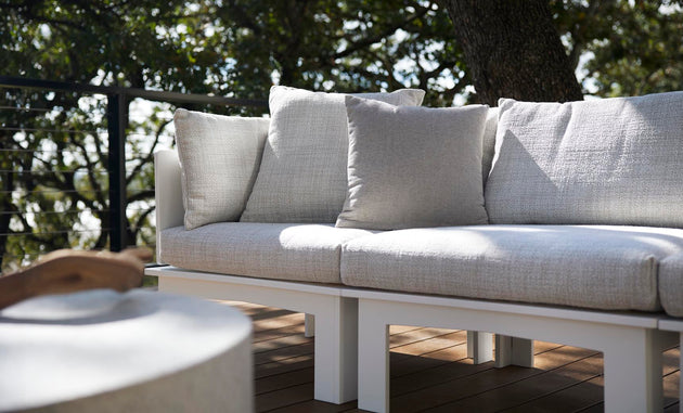 On a wooden deck, a light gray Mainstay Sectional Relaxed Right Corner by Ledge Lounger with cushion set is displayed. A tree with lush green leaves enhances the scene, while a round table is partially visible in the foreground.