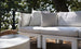 The outdoor patio features a Mainstay Sectional Relaxed Left Armchair with white cushions and light gray pillows on a wooden deck. Metal railings surround the space, with lush trees nearby and a round, light-colored table enhancing the view.